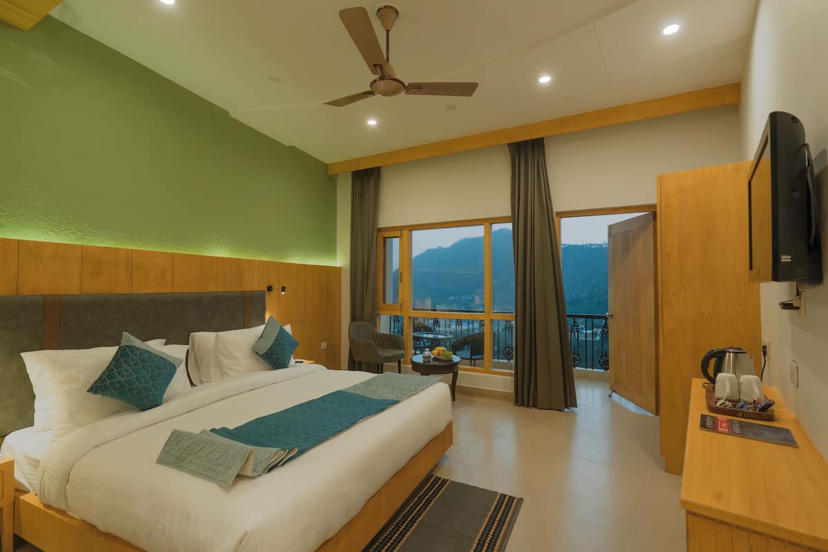 A hotel room with a large bed, a ceiling fan, a wooden headboard, a nightstand, a desk with a coffee maker, and a view of the mountains through large windows.
