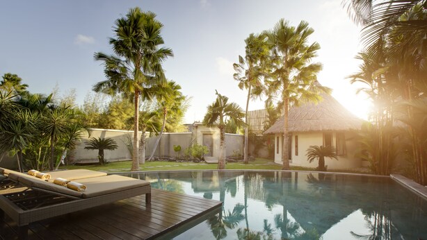 A pool with a wooden deck and lounge chairs, surrounded by palm trees and a thatched-roof building. - Villa Seminyak 3 Bedroom (Seminyak)