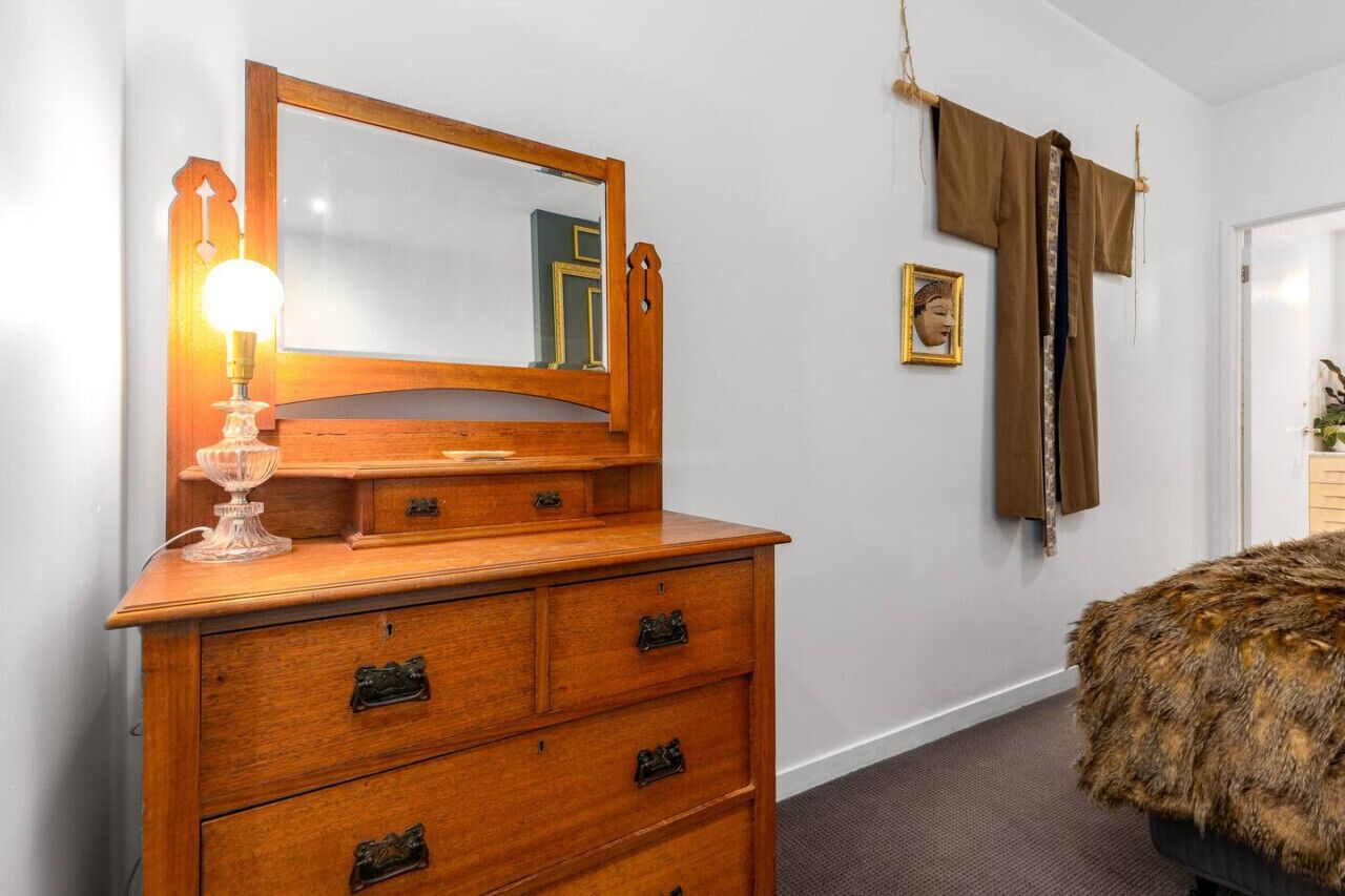 A wooden dresser with a mirror and a light, a fur-covered ottoman, and a brown curtain on a white wall.