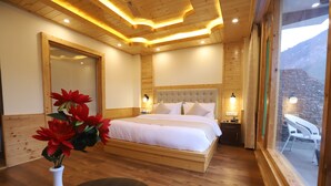 A bedroom with a wooden ceiling, a large bed, a nightstand, a vase with flowers, and a view of the outdoors. - Golden Wood Manali By Rosetum (Manali)