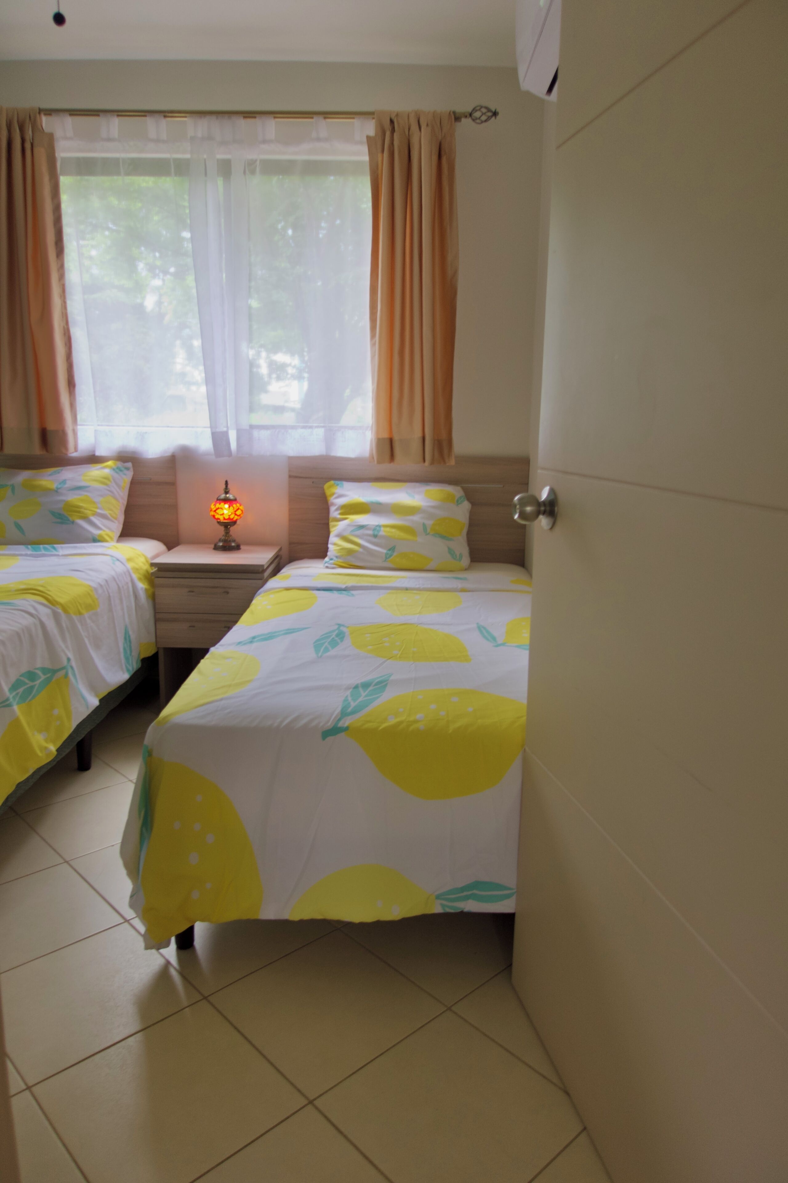 A hotel room with two beds, a nightstand with a lamp, and a window with sheer curtains.