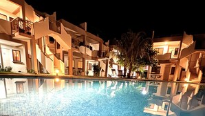 Vista notturna di una piscina alberghiera con pavimento piastrellato e edifici circostanti.