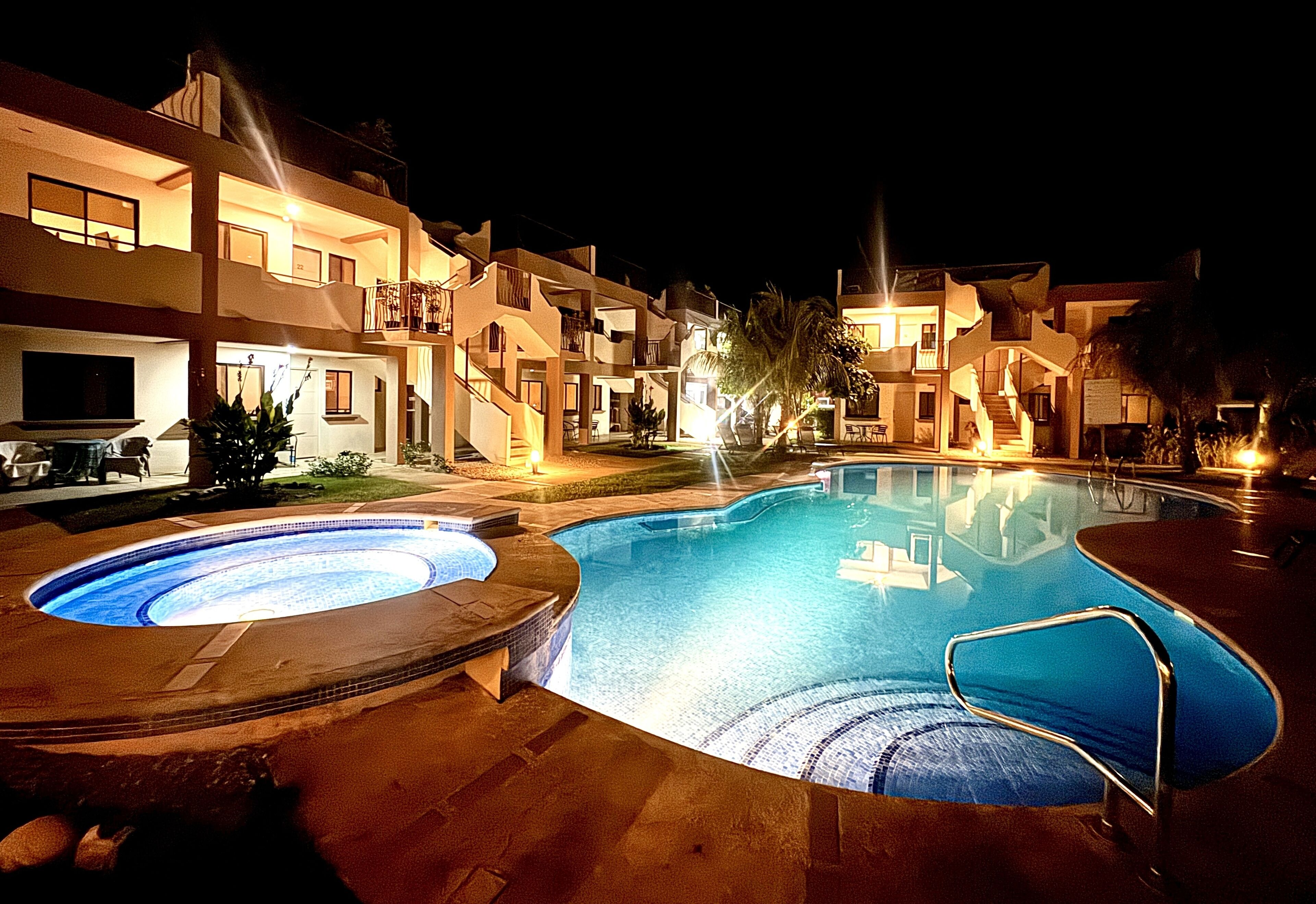 A swimming pool surrounded by multi-story buildings with balconies, a hot tub, and a well-lit outdoor area at night.