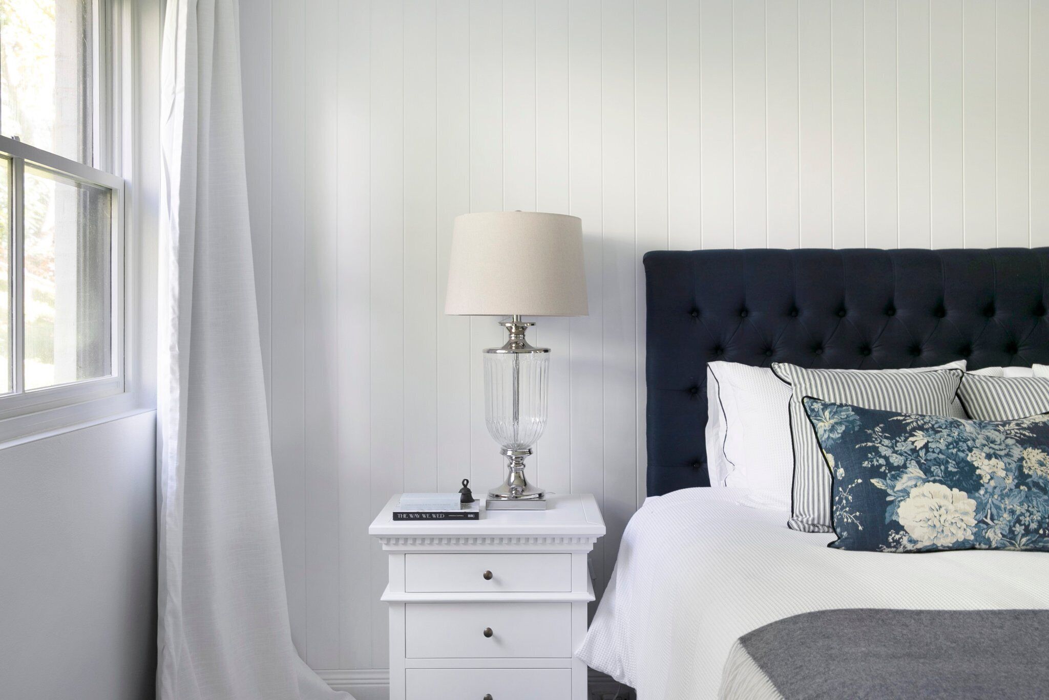 A bedroom with a white nightstand, a lamp, a bed with a dark headboard, and floral pillows.
