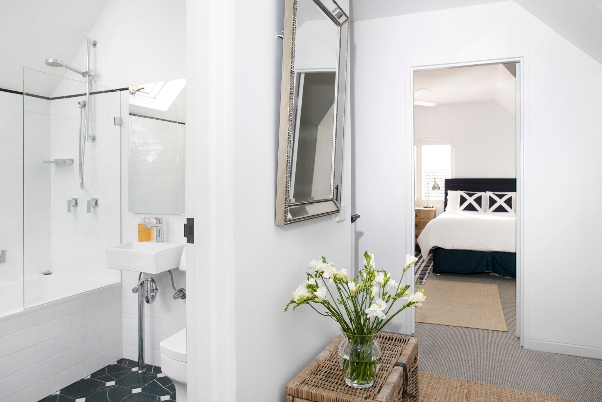 Un baño moderno con cabina de ducha de vidrio, un lavabo blanco y un espejo. Un florero con flores blancas está sobre una mesita auxiliar junto al lavabo.
