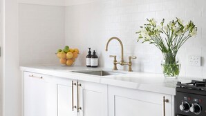 A kitchen with white cabinets, a stainless steel sink, brass faucets, a bowl of fruit, and a vase with green plants.