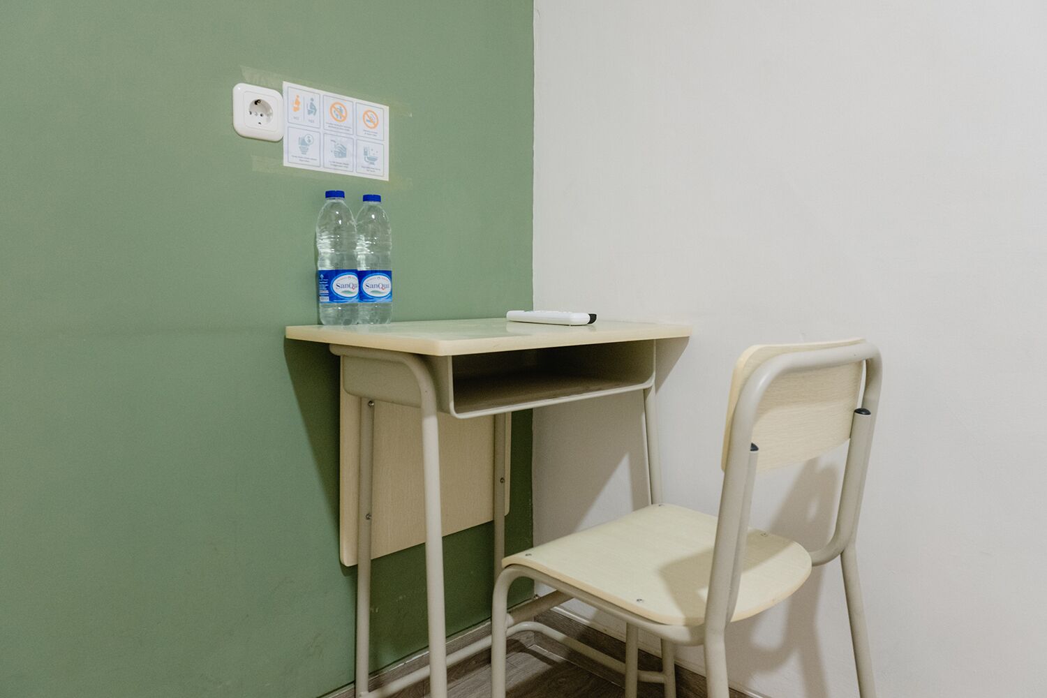 Una habitación pequeña, para una sola persona, con una pared verde, una silla blanca, un escritorio con una computadora y dos botellas de agua.