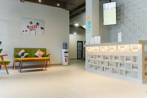 Zona de recepción con un sofá verde, mesa de madera, dispensador de agua y un mostrador blanco con diseño de cuadrícula.