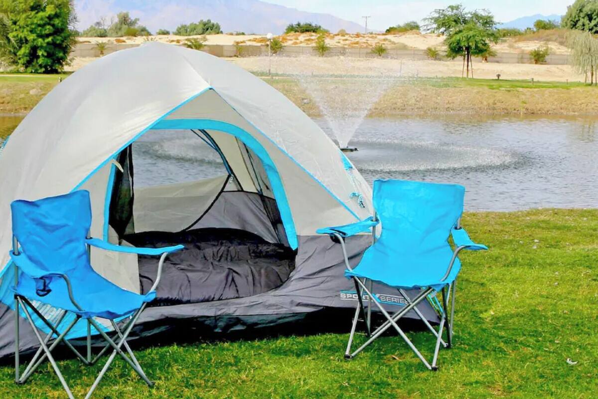 Una tienda de campaña con diseño azul y gris, dos sillas de camping azules, y un lago al fondo.