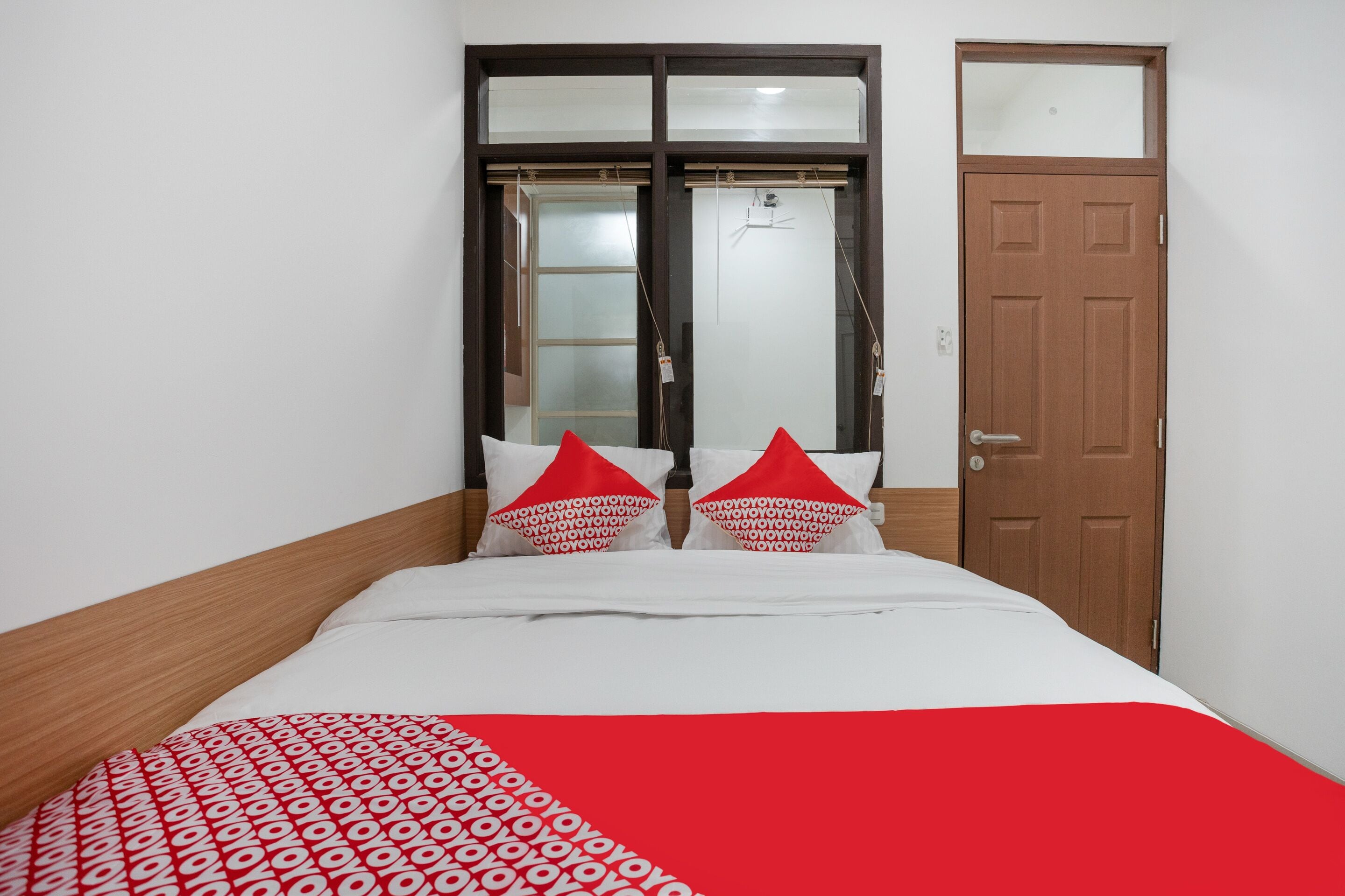 A neatly made bed with a patterned bedspread and two red pillows, a wooden headboard, a window with blinds, and a door.