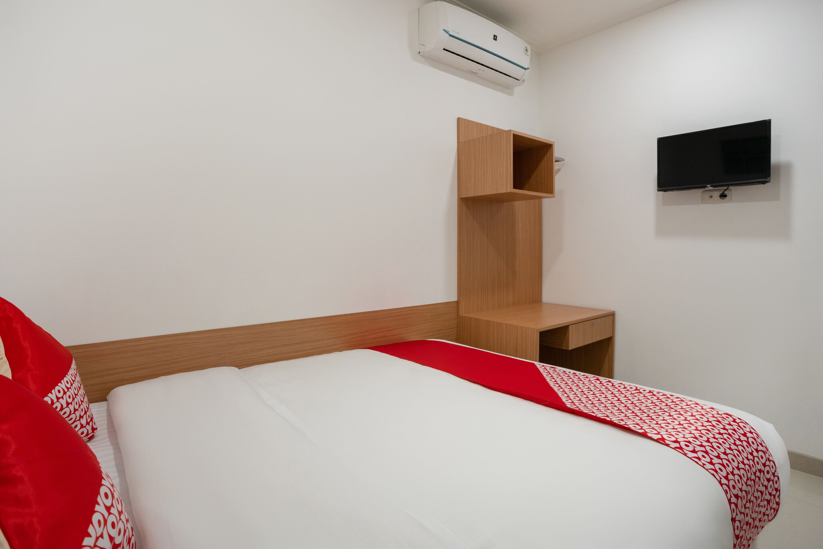 A hotel room with a bed, a wooden nightstand, a TV mounted on the wall, and an air conditioning unit.