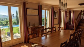 A dining area with a large wooden table and chairs, a view of a pool and greenery through glass doors, and a staircase leading to an upper level. - Large House with Private Pool and Valley View (Savigny-sur-Grosne)