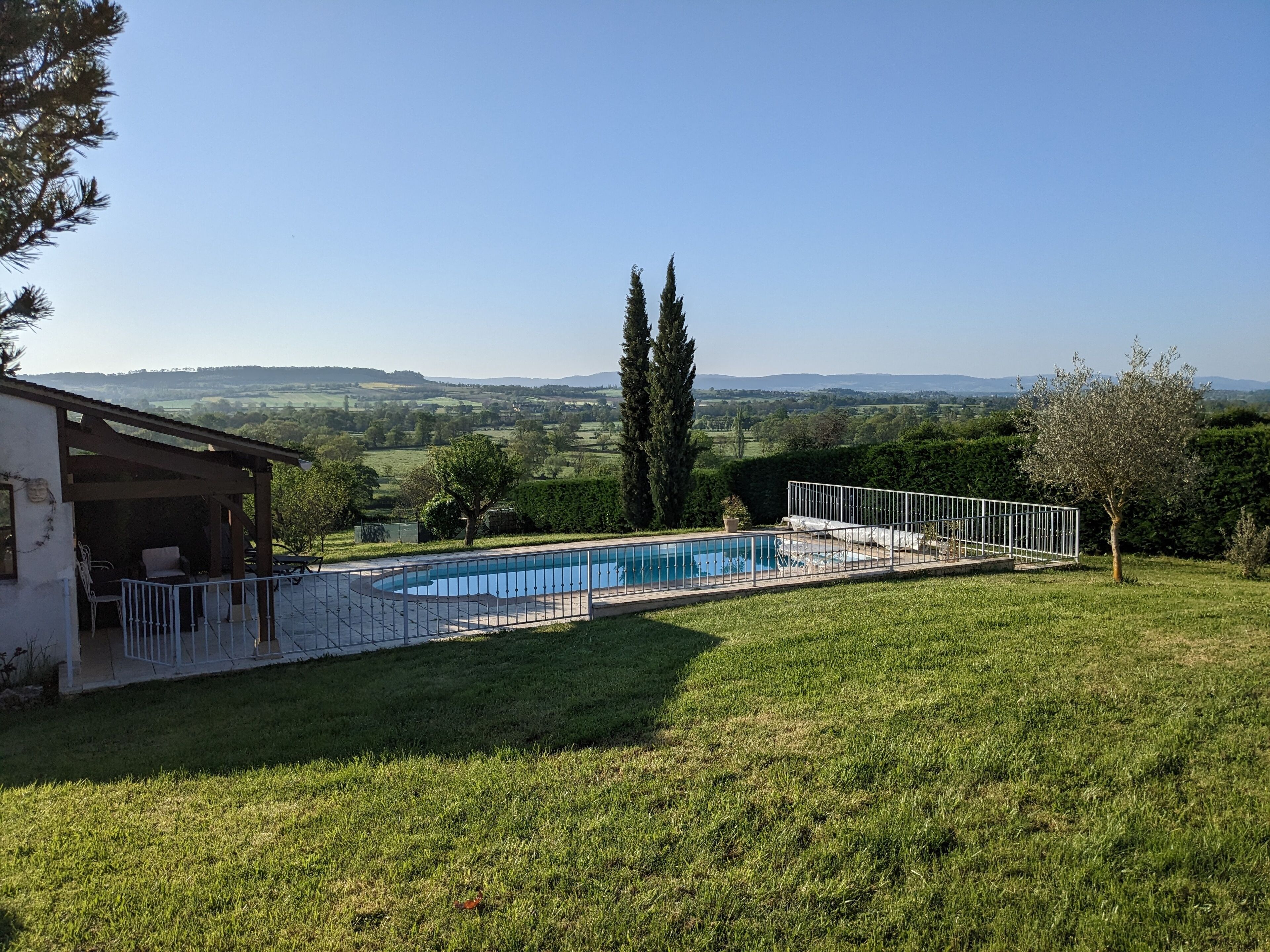 A swimming pool with a white fence, surrounded by a grassy lawn and trees, with a view of distant hills.