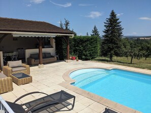 A pool area with a covered seating area, outdoor furniture, and a view of the surrounding landscape.