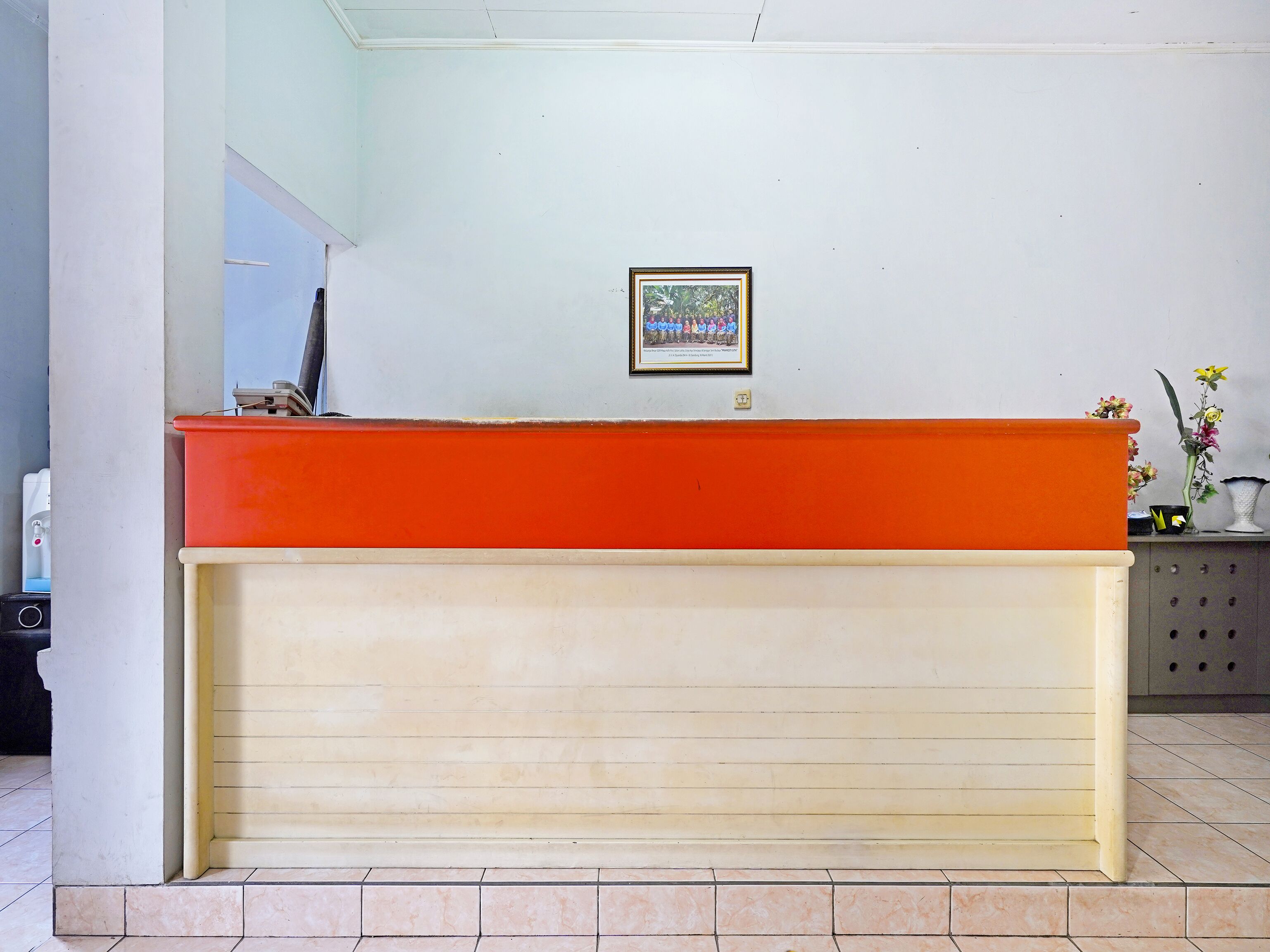 A reception desk with a beige lower section and an orange upper section, a framed picture on the wall, and a small potted plant on the right.