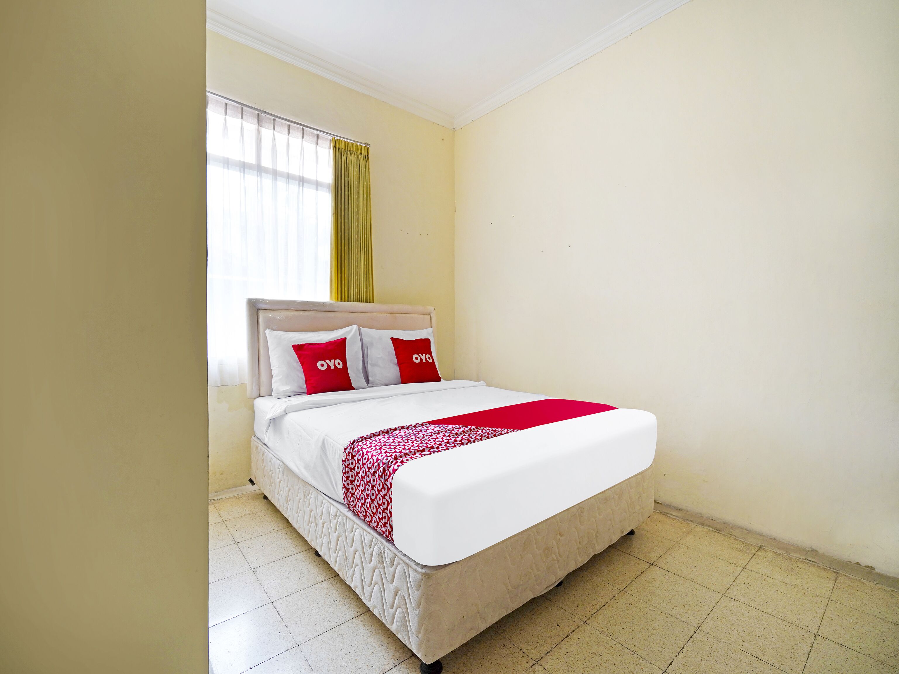 Una cama individual con colcha blanca y roja, dos almohadas con la marca 'OYO', y una ventana con cortinas amarillas.