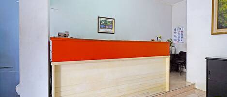 A reception desk with a red top and beige lower section, a framed picture on the wall, a calendar, and a potted plant.