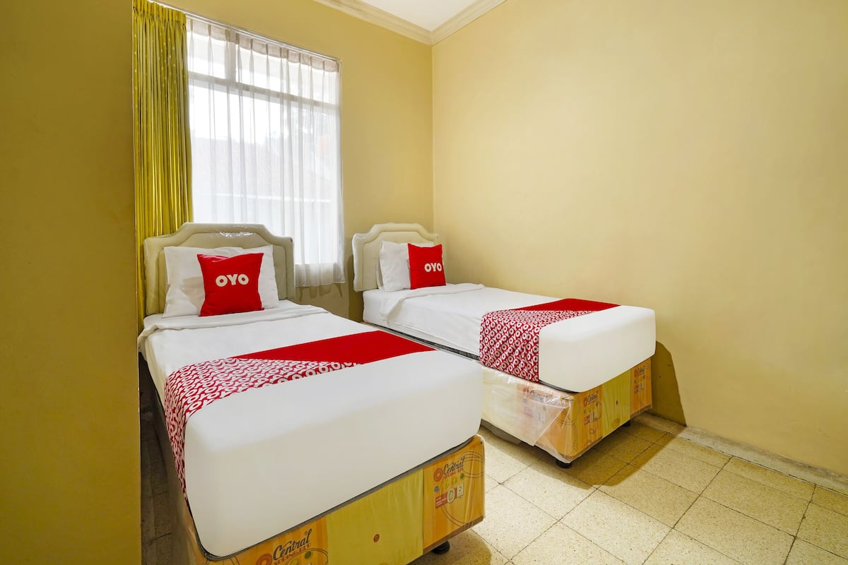 Ein Hotelzimmer mit zwei Betten, jeweils mit weißer Matratze und roter Bettwäsche, und einem Fenster mit gelben Vorhängen.
