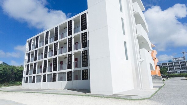 Un bâtiment moderne, blanc, à plusieurs étages et doté de balcons, entouré d’un ciel dégagé et de verdure.