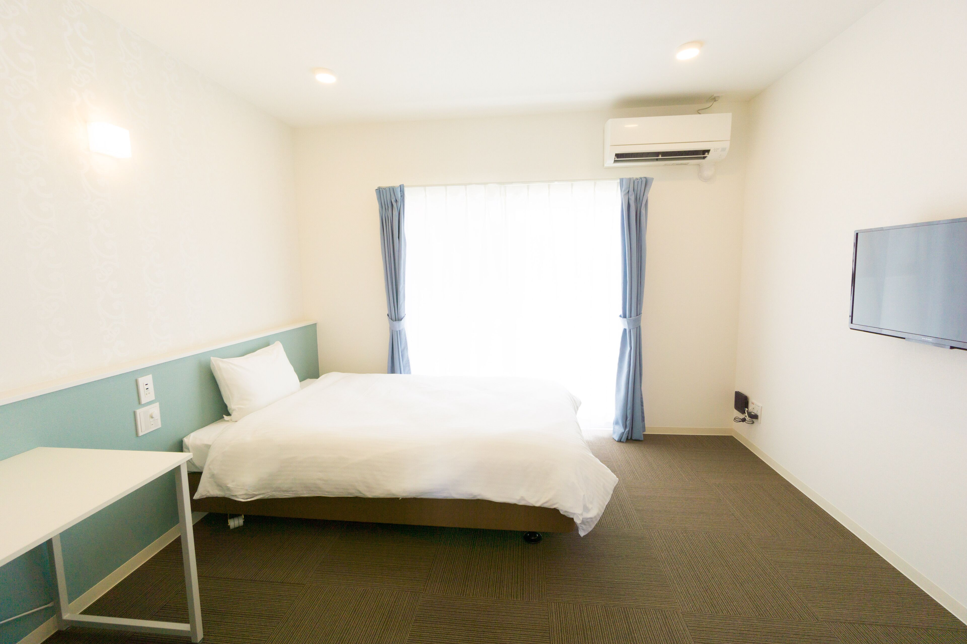 A hotel room with a bed, a desk, a TV, and an air conditioning unit.