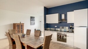 Une cuisine moderne avec des meubles blancs, un four encastré et un coin repas comprenant une table en bois et des chaises en osier.