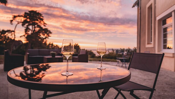 Une table ronde avec deux verres à vin, placée sur une terrasse d’où l’on peut admirer le coucher du soleil.