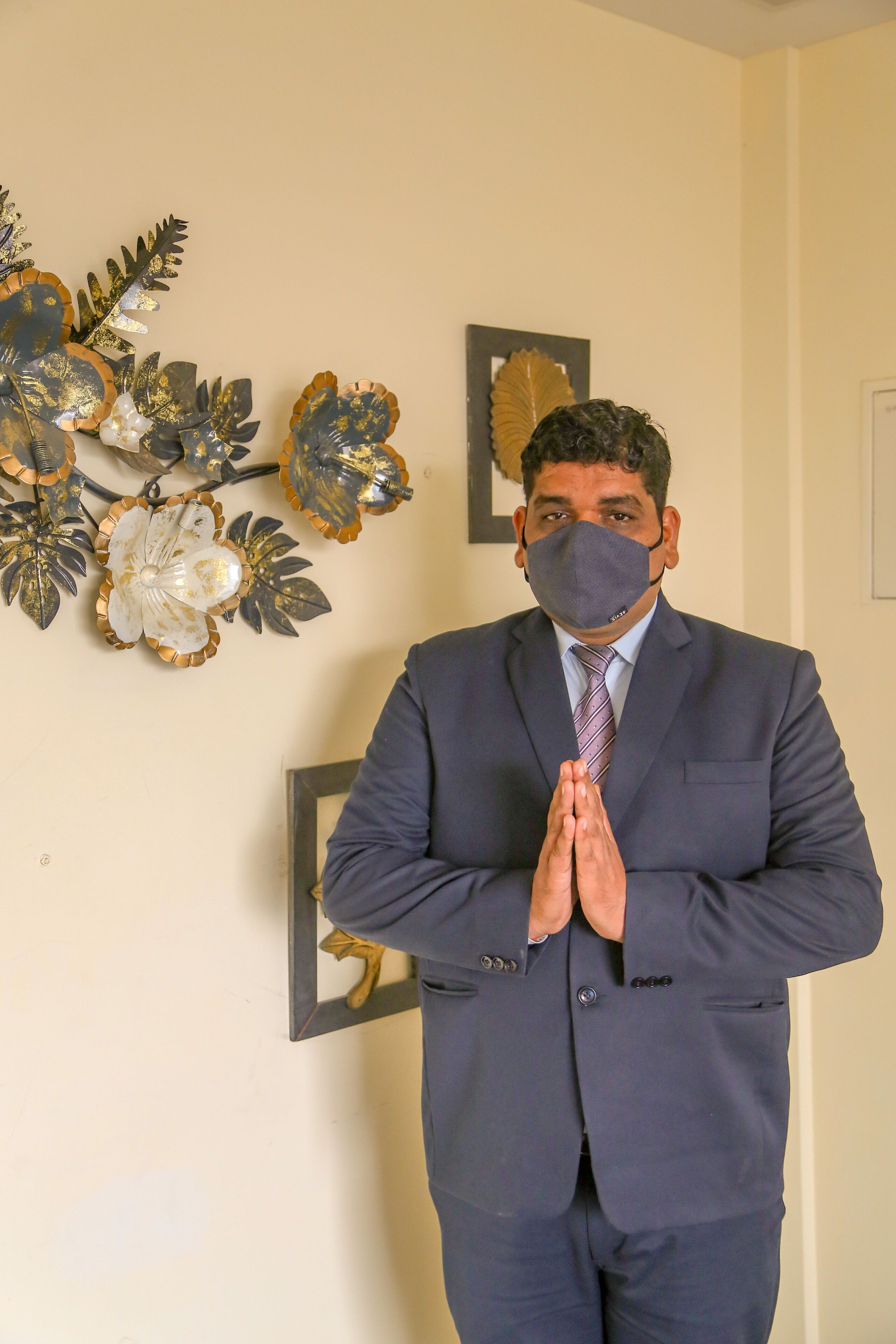 A man in a suit standing in a room with a wall decorated with metallic leaf designs.