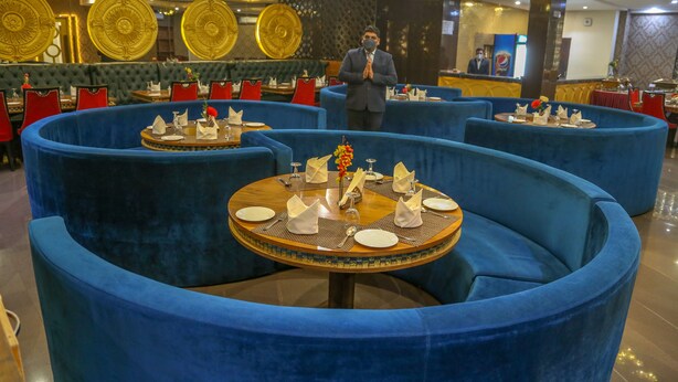 A restaurant with round tables and blue upholstered chairs, a man standing in the background, and a patterned wall. - Four Leaf Hotel (Budaun)