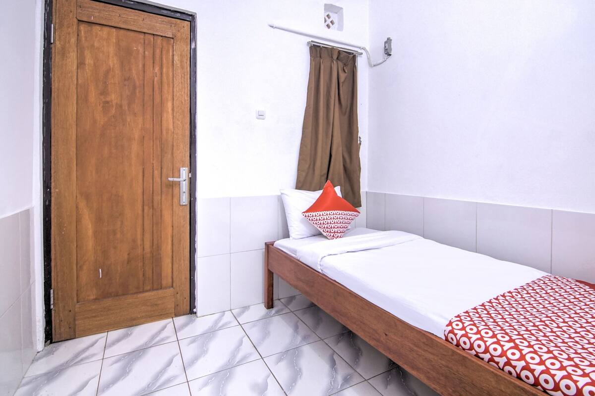 Une petite pièce avec une porte en bois, un lit simple recouvert d’une couverture à motifs, un mur blanc et un rideau marron.