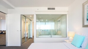 Une chambre d’hôtel moderne dotée d’un grand lit, d’une paroi en verre donnant sur la salle de bain et d’une petite table de chevet avec une lampe.