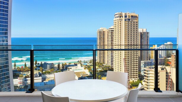 Un balcon avec une table et des chaises blanches, donnant sur une plage et l’océan, avec des immeubles de grande hauteur en arrière-plan.