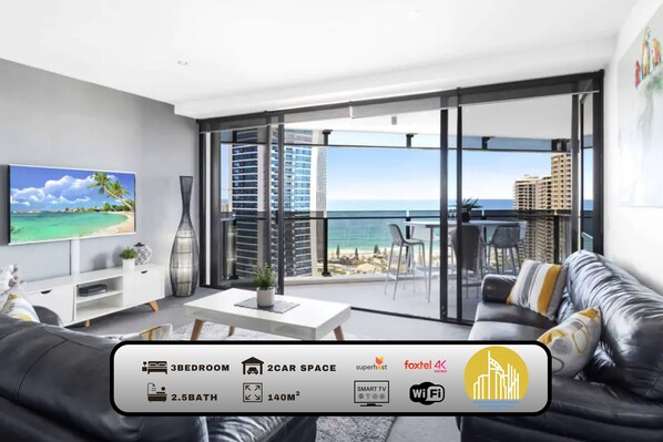Een moderne woonkamer met een grote televisie, een bank en een balkon met uitzicht op de oceaan en gebouwen. Het appartement heeft 3 slaapkamers, 2 parkeerplaatsen, 2,5 badkamers, 140 vierkante meter, een smart-tv en WiFi.