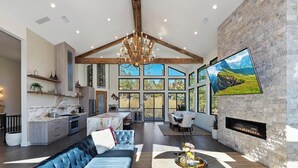 A spacious living area with a high ceiling, a large chandelier, a fireplace, and a dining area with a view through large windows. - Bella Villa: Luxury On Bear Mountain Golf Course! Air Conditioning! Spa! Theatre! Game Room! Walk to slopes! (Big Bear Lake)