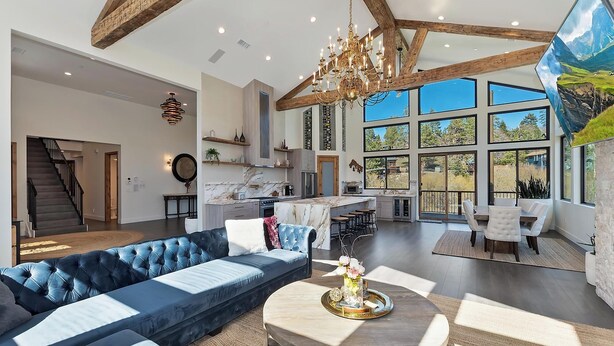 A spacious living area with a large sofa, a round coffee table, and a view of the kitchen and outdoor scenery through large windows. - Bella Villa: Luxury On Bear Mountain Golf Course! Air Conditioning! Spa! Theatre! Game Room! Walk to slopes! (Big Bear Lake)
