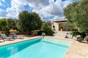 Piscina com espreguiçadeiras, cercada por árvores e um edifício com degraus que levam a uma porta.