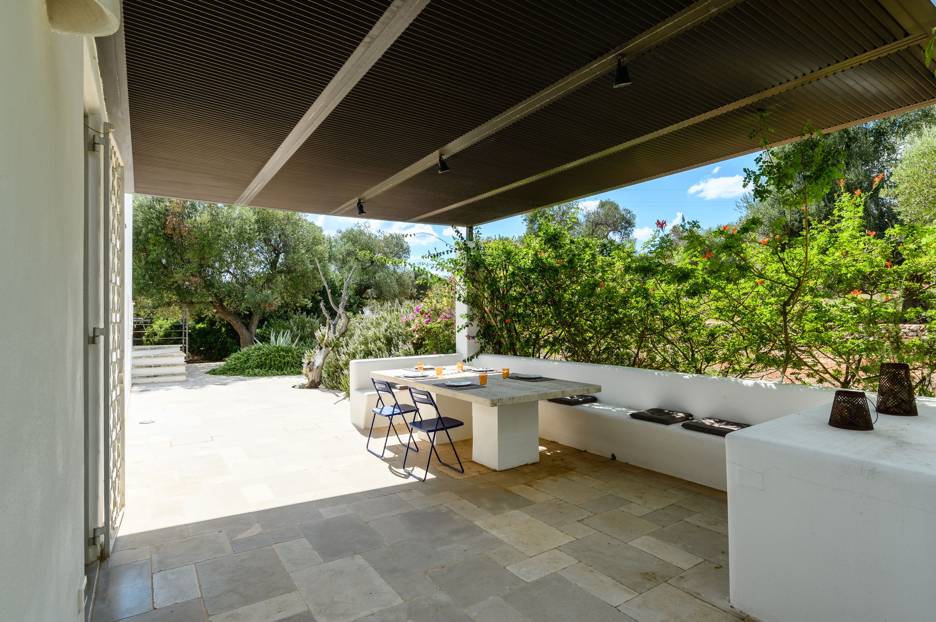 An outdoor patio with a table and chairs, surrounded by greenery and a white wall.