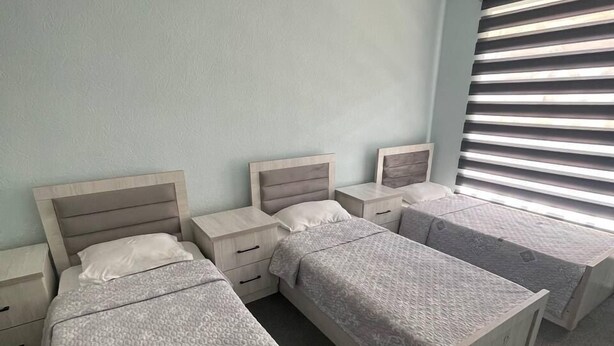 A room with three single beds, each with a wooden headboard and a grey patterned bedspread. There are bedside tables with drawers and a window with horizontal blinds.