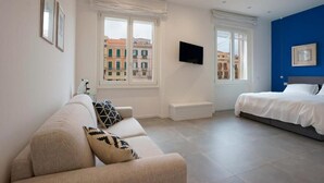 A modern bedroom with a sofa, a bed, a TV, and large windows offering a city view. - Suite nel Largo (Cagliari)