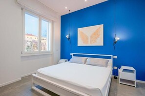 Une chambre avec un lit blanc, un mur d’accent bleu et une fenêtre donnant sur des immeubles.