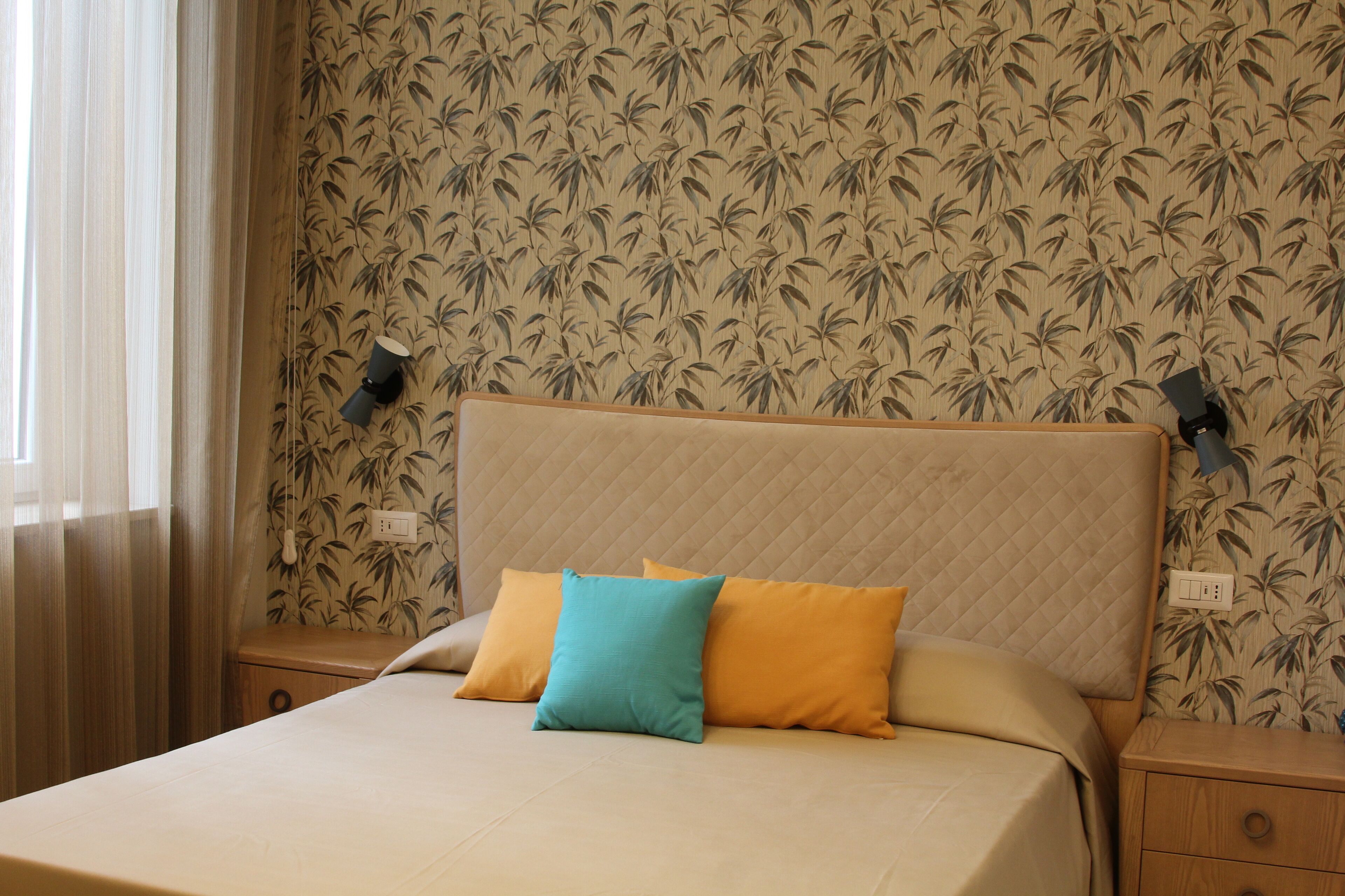 A neatly made bed with a quilted headboard and two pillows, a nightstand with a lamp, and a wall with a bamboo leaf pattern.