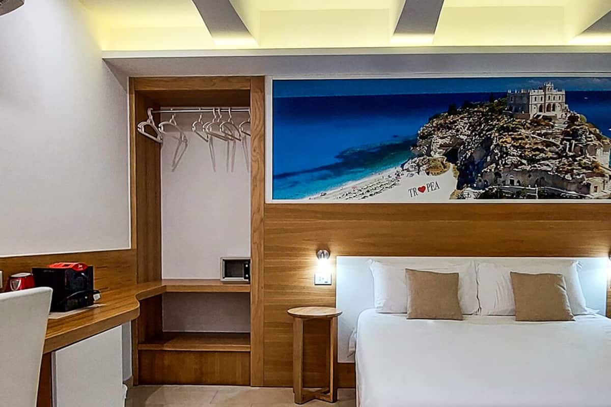 A hotel room with a bed, a desk, a chair, a small table, and a painting of a coastal scene.