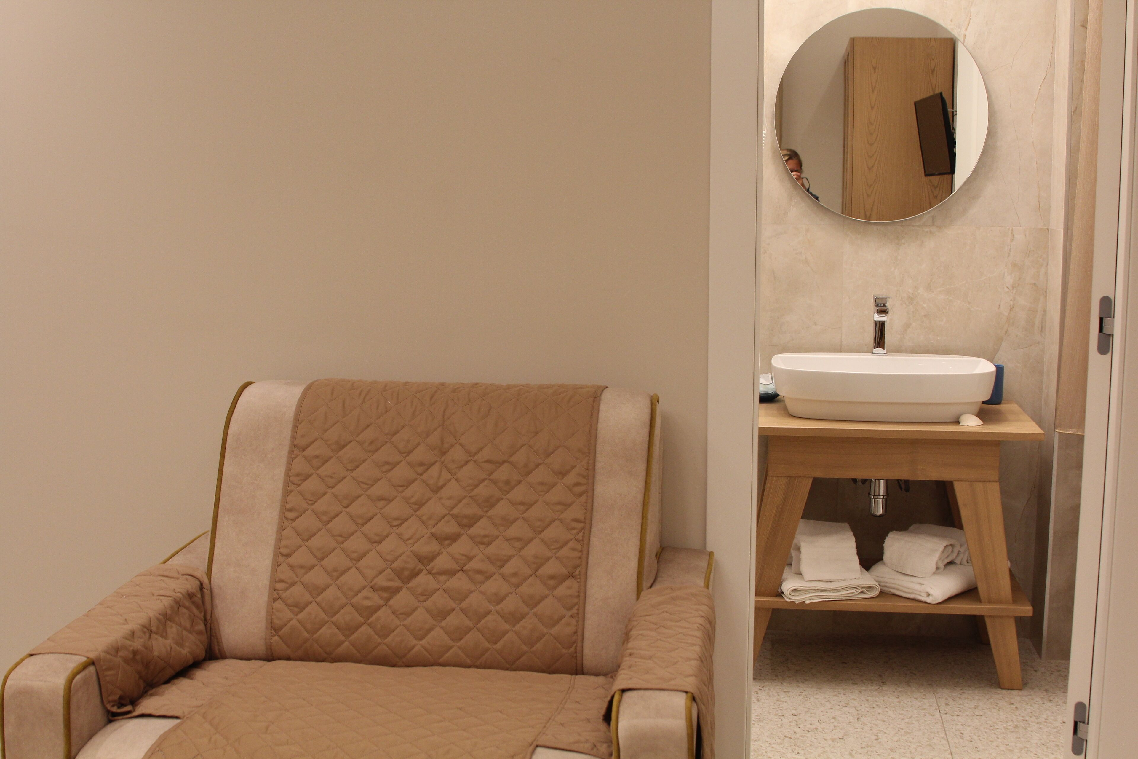 Un baño moderno con un sillón color beige, un espejo redondo y un tocador de madera con lavamanos y espacio para toallas.