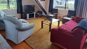 Sala de estar com sofá, poltronas, mesa de centro e televisão. Possui janelas com cortinas, teto de madeira e vista para uma casa e colinas.