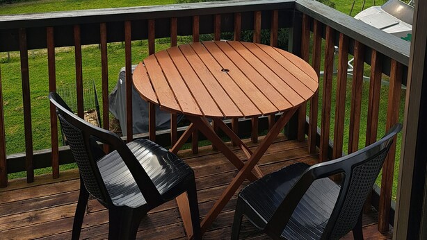 Um deck de madeira com uma mesa redonda e duas cadeiras, com vista para uma área residencial com casas e o mar.