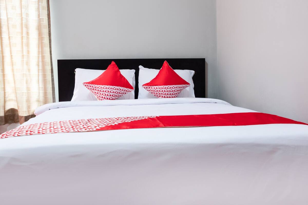 Una cama bien hecha con ropa de cama blanca y roja, un cabecero negro y una ventana con cortinas.