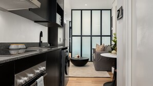 A modern kitchen with a built-in oven, a black stove, and a sink. There is a round black table with a bowl and chopsticks. A gray sofa and a small round table with a plant are in the background.