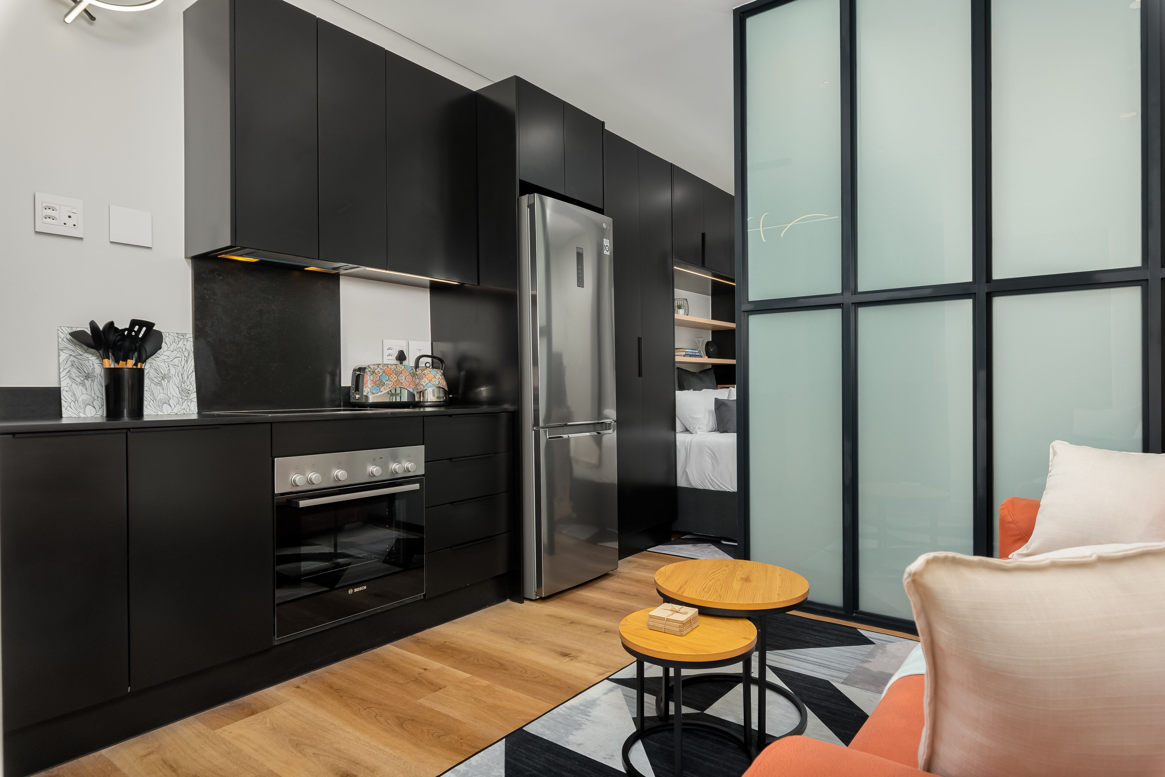 Una cocina moderna con armarios negros, un refrigerador de acero inoxidable y un horno empotrado. Hay una pequeña mesa de madera con dos taburetes y un sofá con cojines naranjas.