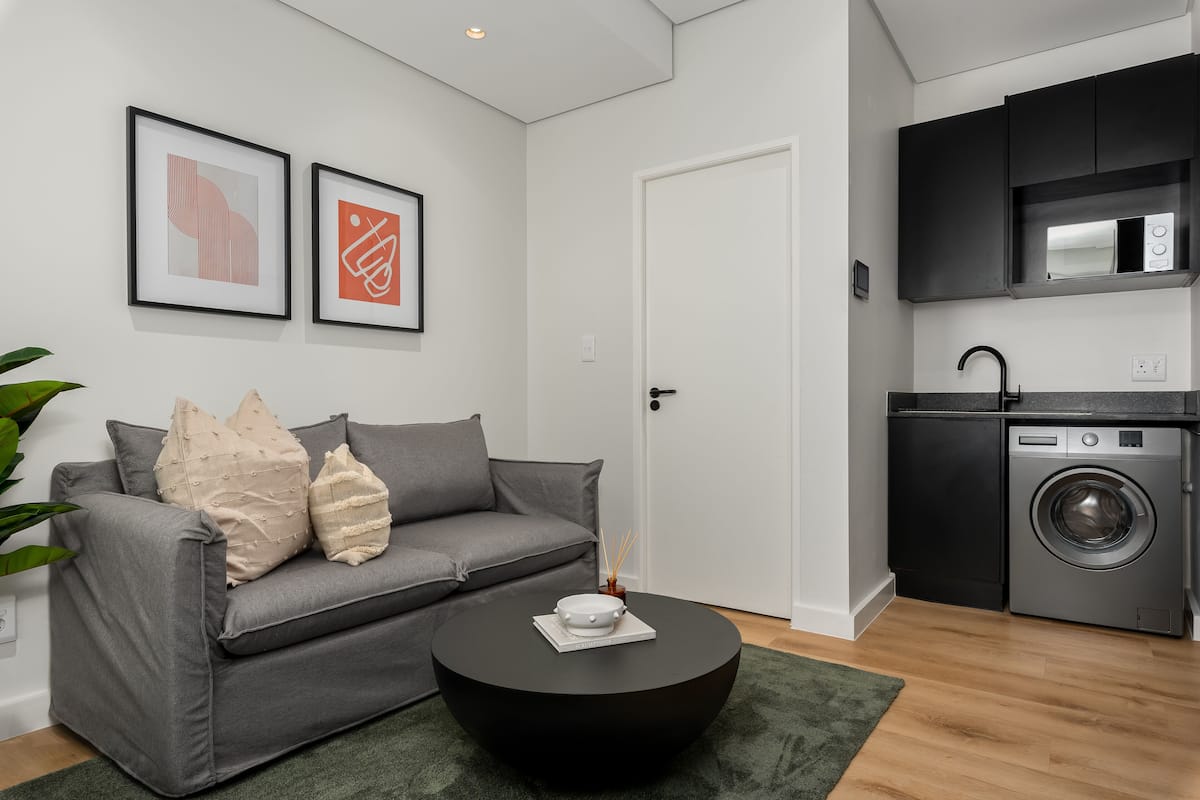 Una sala de estar moderna con un sofá gris, una mesa de centro negra y una alfombra verde. Hay una cocina con lavadora y armarios.