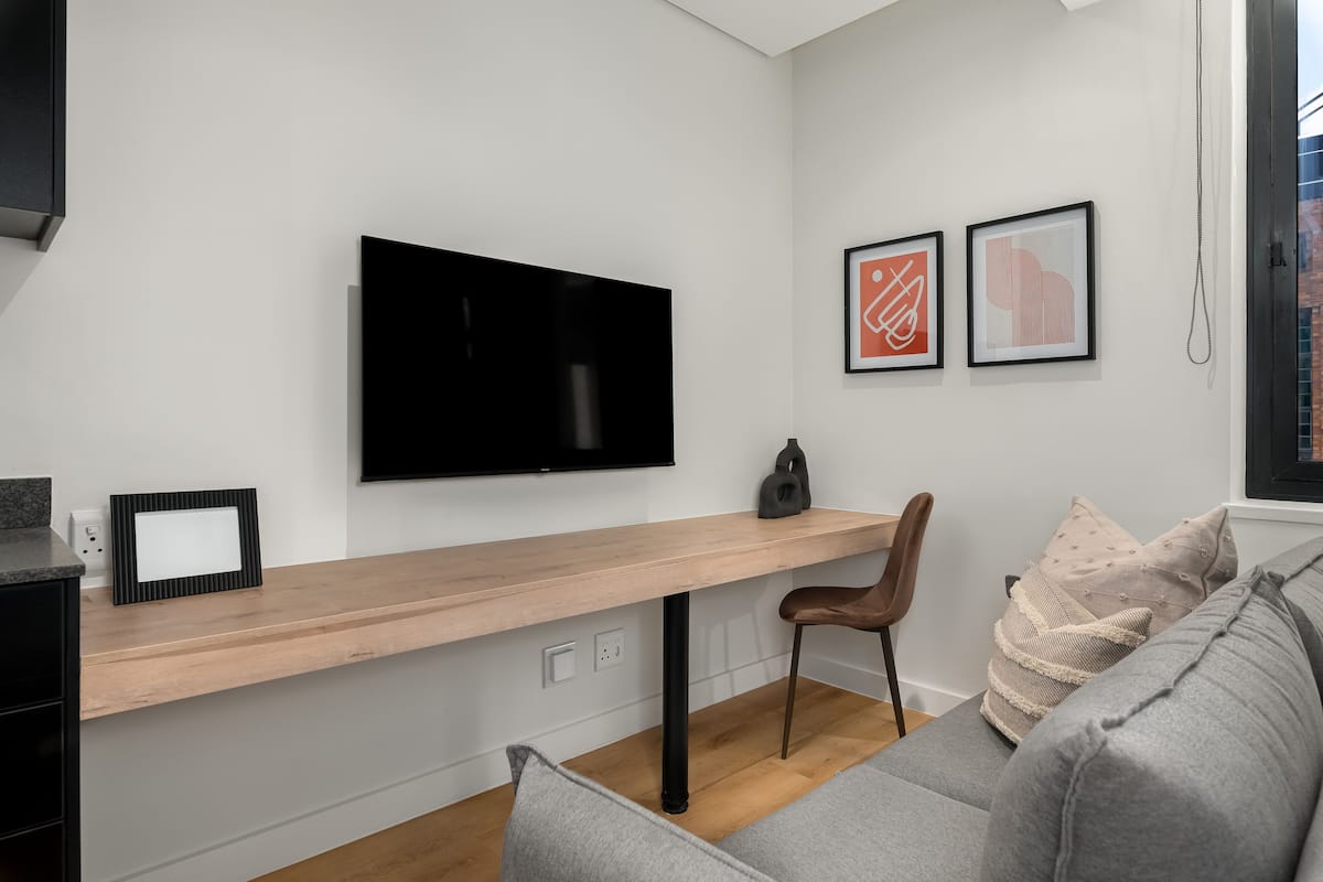 Una sala de estar moderna con un televisor de pantalla plana montado en la pared, un escritorio de madera, un sofá gris y dos obras de arte abstracto enmarcadas.