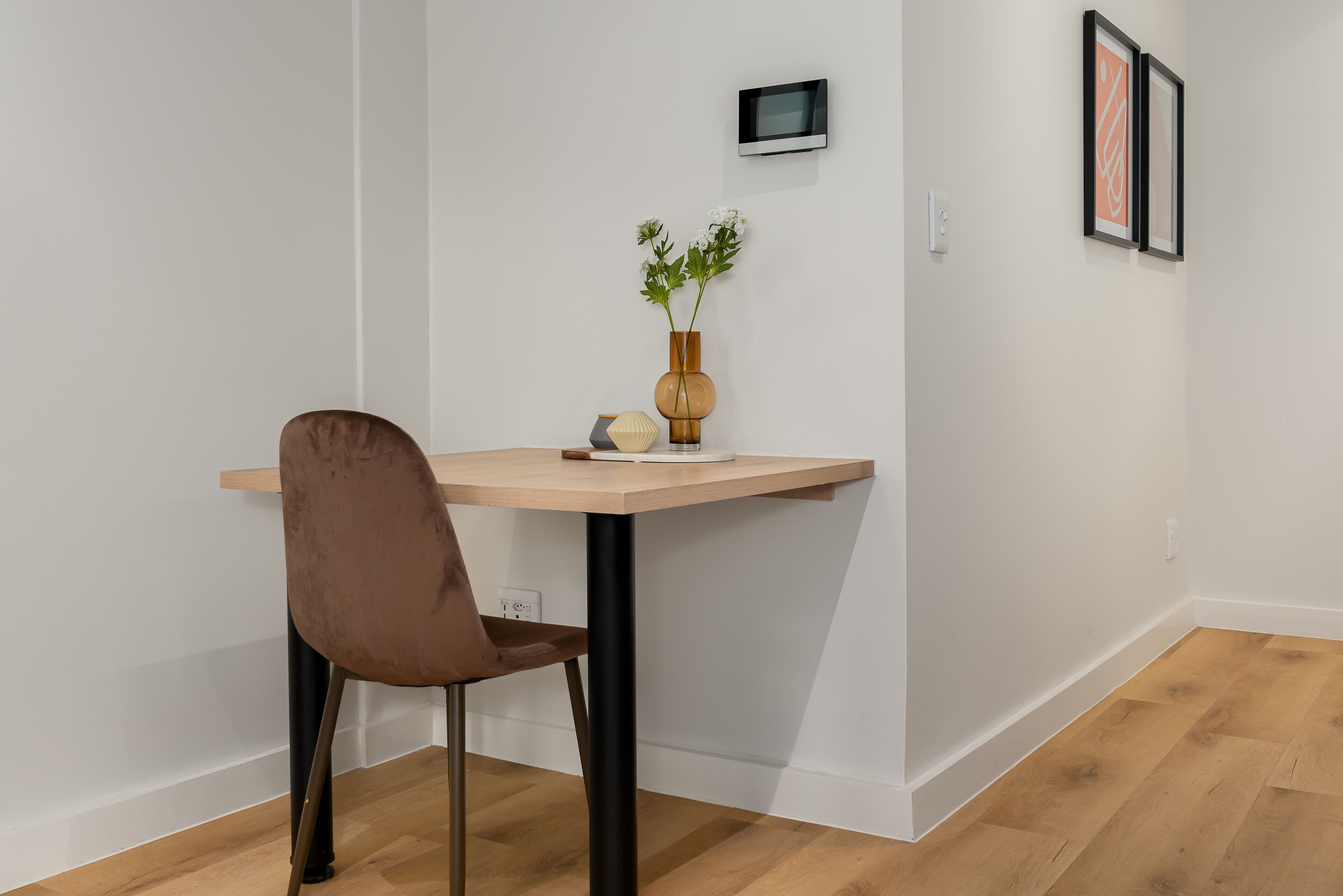 A modern room with a wooden desk, a brown chair, a vase with flowers, and framed artwork on the wall.